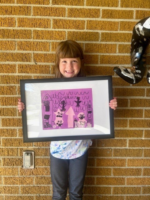 Smiling student with dark hair holding a framed picture of her mix-media artwork that features two houses, shape people, and lots of hand drawn birds.