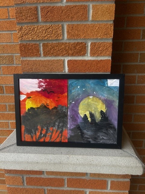 Photograph of a framed painting against a brick building. Painting features a sunrise landscape on one side and a large moon landscape on the other side.
