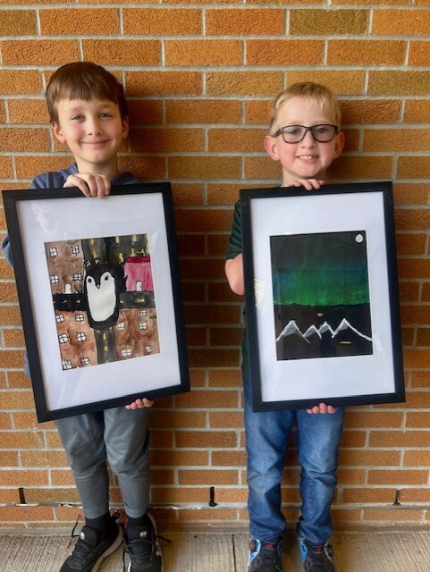 Two students smiling and holding their framed artworks. First student has dark hair and has a picture of a penguin in a city scene. Blond student has a painting of the Northern lights on a mountain range.
