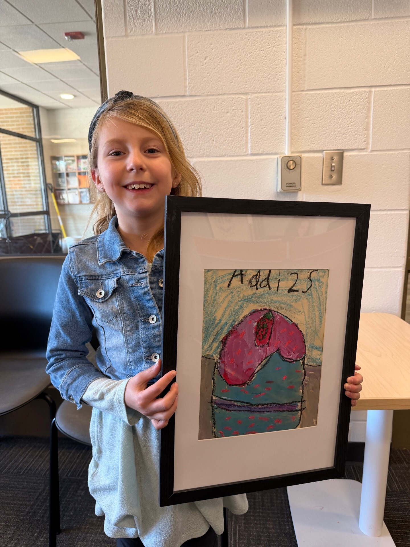 Students with long blond hair poses behind her framed artwork of a blue and purple cupcake.
