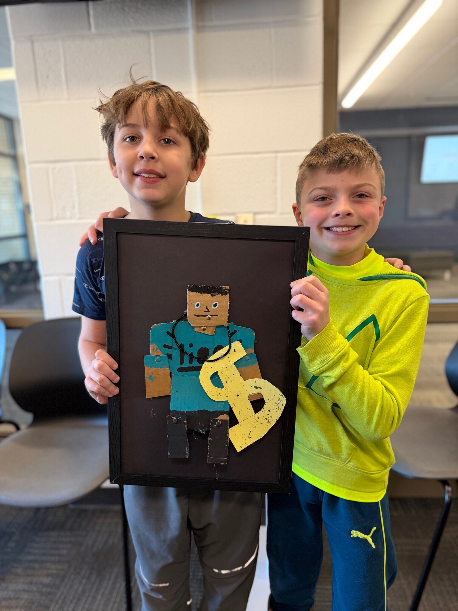 Image of two smiling students standing behind their framed artwork with a cardboard man wearing a large dollar sign necklace.