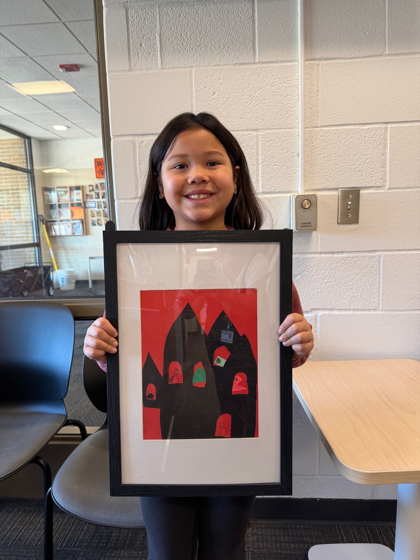 Smiling students with shoulder length dark hair poses behind her framed artwork. A collage of black construction paper houses on a red background.
