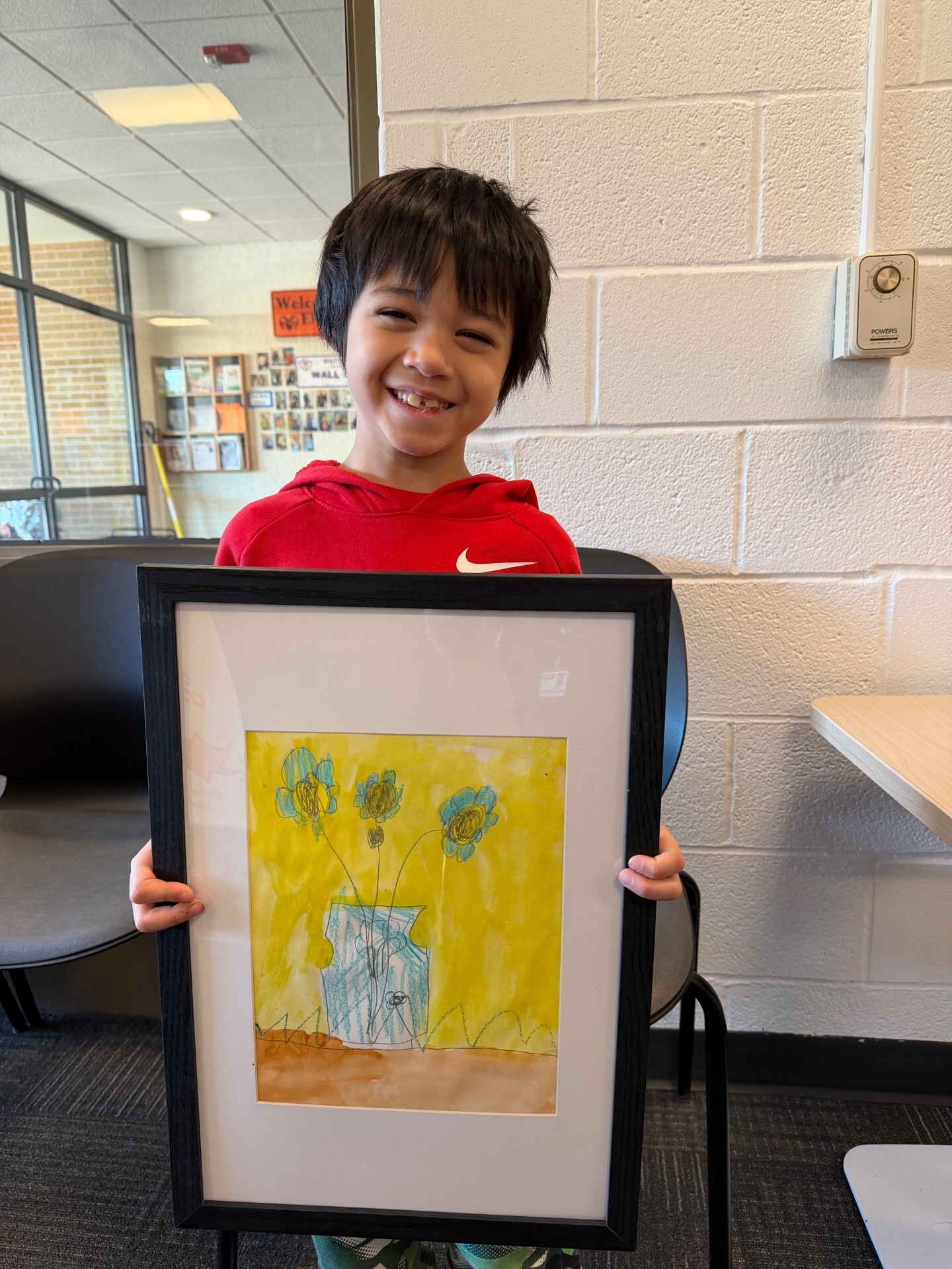 Smiling student with dark hair and a red sweatshirt poses behind his artwork that features a vase of flower on a yellow background.