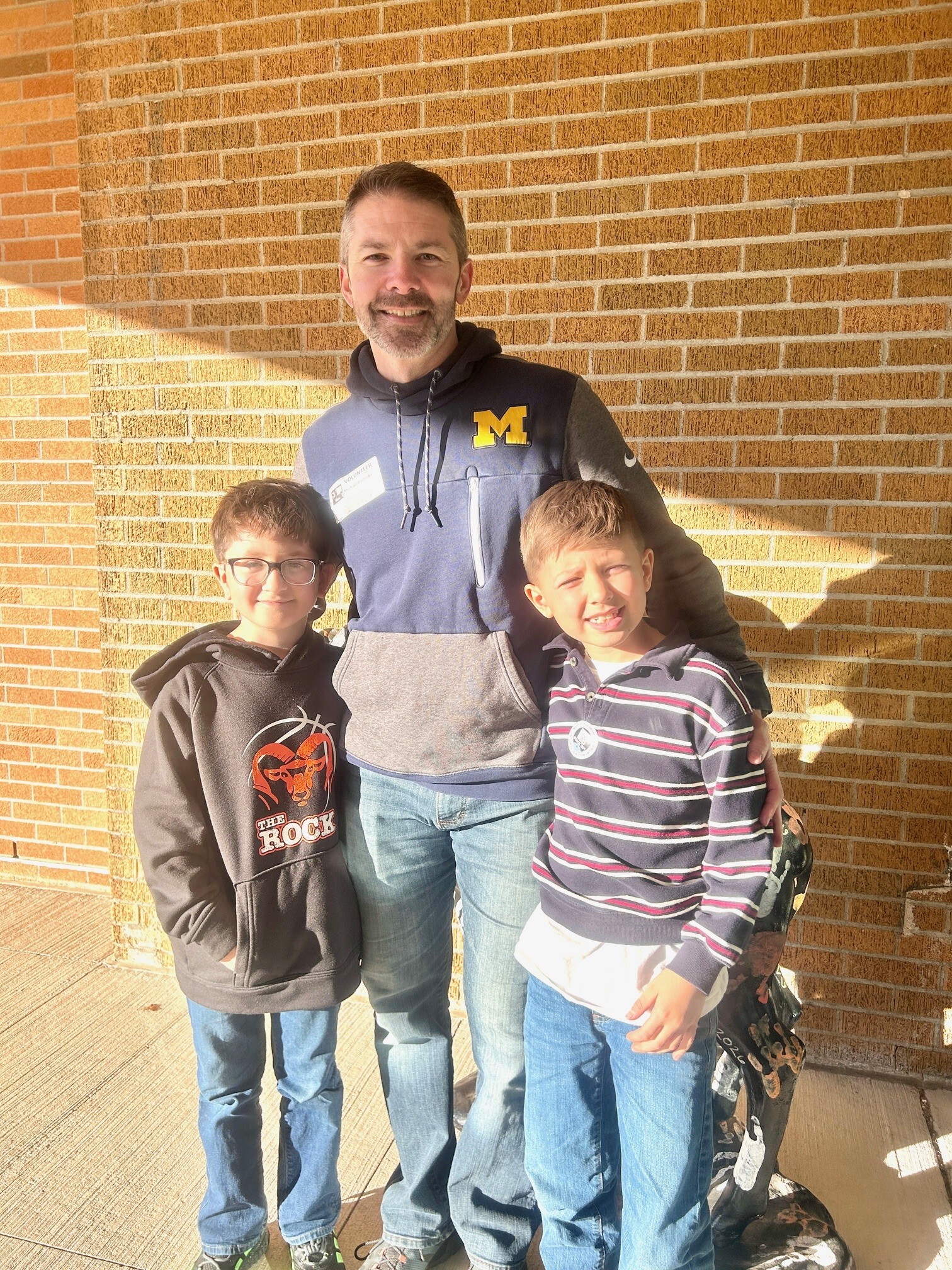 A man with a Michigan sweatshirt stands with his two smiling sons in front of a brick wall.